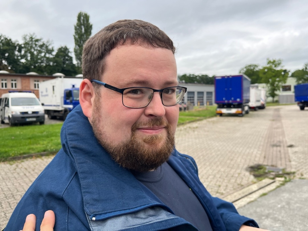 Johannes Rudolph in blauer THW-Einsatzkleidung auf dem Gelände eines THW-Ortsverbands, dahinter Fahrzeuge und Anhänger.